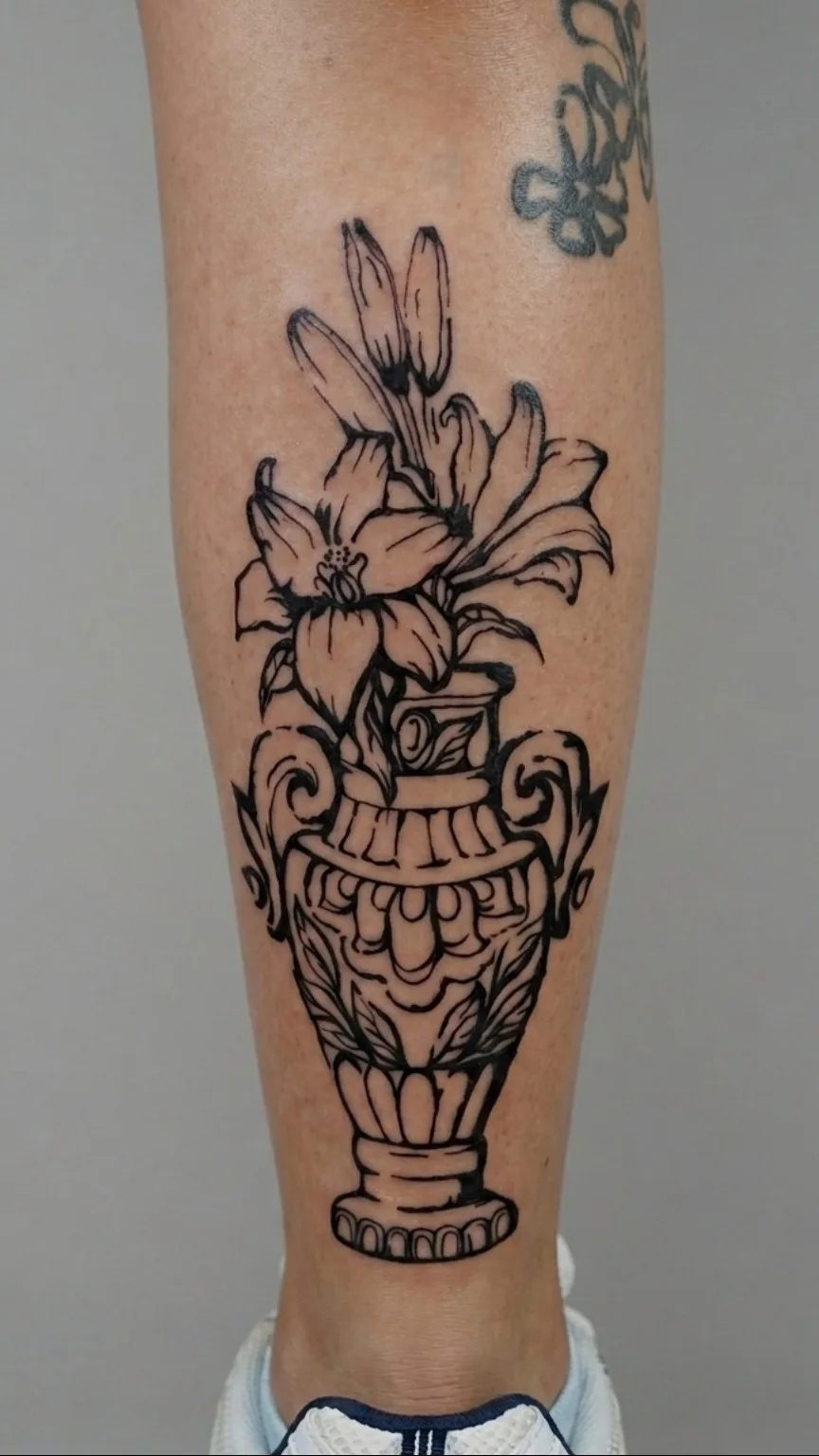 Tatuaje de un jarrón ornamental con flores y hojas en tinta negra, sobre la pantorrilla.