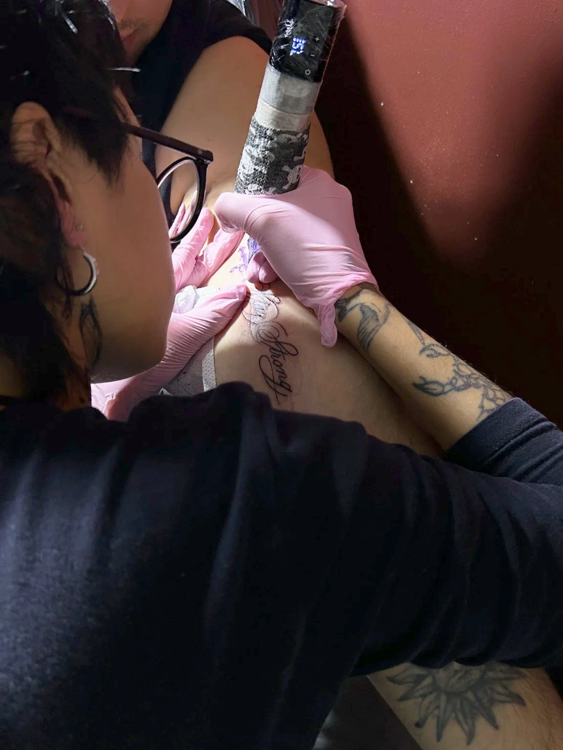 Nat, con guantes rosados, tatúa un lettering con la máquina en la mano.