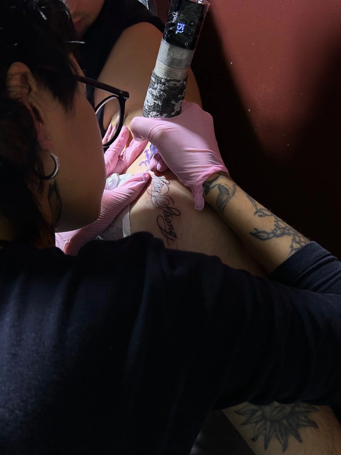 Nat, con guantes rosados, tatúa un lettering en el hombro de un cliente.